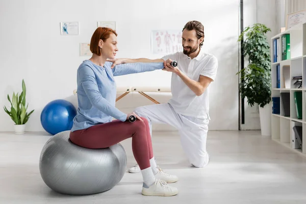 Fisioterapista sorridente che aiuta la donna rossa seduta sulla palla fitness e che lavora con i manubri nella clinica di riabilitazione — Foto stock