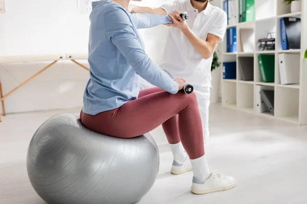 Обрезанный вид женщины, сидящей на фитболе и упражняющейся с гантелями возле физиотерапевта в реабилитационном центре — стоковое фото