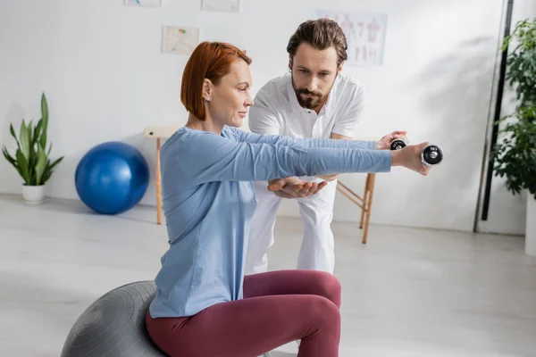Bärtige Physiotherapeutin unterstützt rothaarige Frau, die auf Fitball sitzt und im Reha-Zentrum mit Hanteln trainiert — Stockfoto