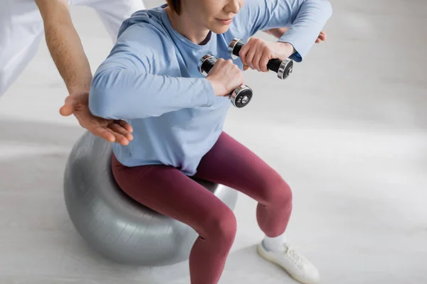 Vista parcial de la mujer sentada en la pelota de fitness y haciendo ejercicio con pesas cerca de rehabilitólogo en la clínica - foto de stock