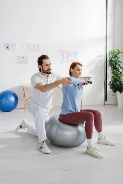 Especialista en rehabilitación asistiendo a una mujer sentada en una pelota de fitness y haciendo ejercicio con pesas en el hospital - foto de stock