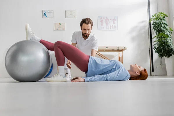 Frau liegt am Boden und trainiert mit Fitball in der Nähe des bärtigen Physiotherapeuten im Reha-Zentrum — Stockfoto