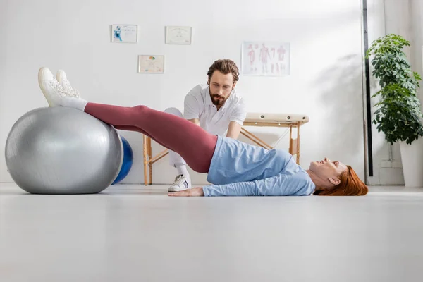Mulher ruiva treinando com bola de fitness enquanto deitado perto fisioterapeuta no centro de reabilitação — Fotografia de Stock
