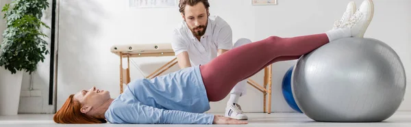Бородатий фізіотерапевт дивиться на жінку, що лежить на підлозі і працює з фітболом у центрі реабілітації, банер — стокове фото