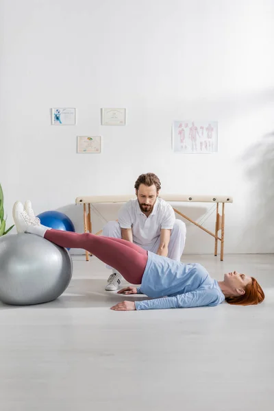 Bärtige Physiotherapeutin neben Frau, die am Boden liegt und im Reha-Zentrum mit Fitnessball trainiert — Stockfoto