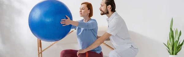 Fisioterapista barbuto che assiste la donna che si allena con la palla fitness nel centro di recupero, banner — Foto stock