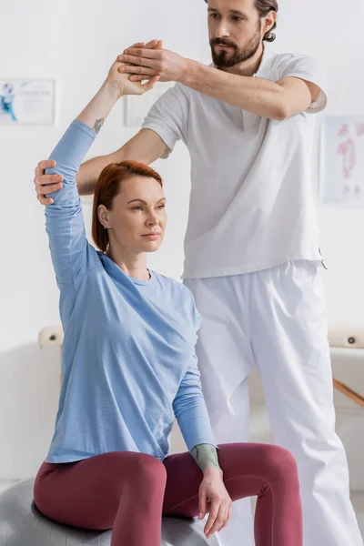 Physiotherapist supporting arm of redhead woman training on fitness ball in recovery clinic — Stock Photo