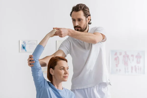 Fisioterapeuta barbudo brazo de apoyo de mujer pelirroja haciendo ejercicio en el centro de rehabilitación - foto de stock