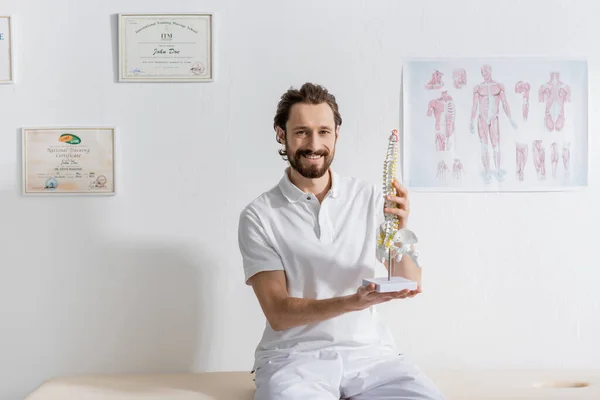 Fröhlicher Chiropraktiker lächelt in die Kamera, während er mit Wirbelsäulenmodell im Reha-Zentrum sitzt — Stockfoto