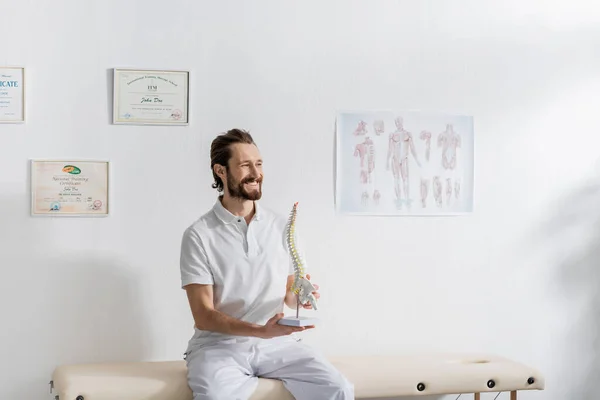 Fisioterapeuta alegre con modelo de columna vertebral mirando hacia otro lado mientras está sentado en la mesa de masaje en el centro de rehabilitación - foto de stock