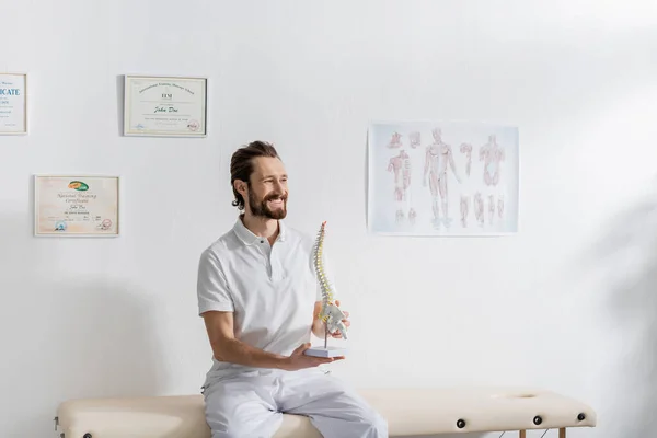 Cheerful physiotherapist with spine model looking away while sitting on massage table in rehabilitation center — Stock Photo