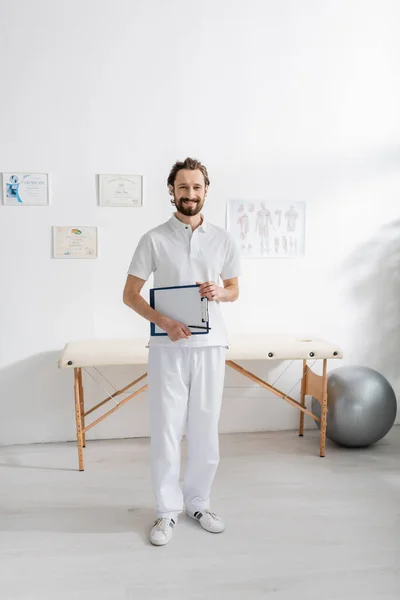 Full length of carefree physiotherapist holding clipboard with paper while standing in consulting room — Stock Photo