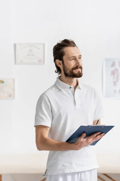 Smiling bearded chiropractor looking away in consulting room of recovery center — Stock Photo