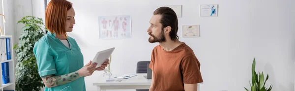 Татуированный физиотерапевт с цифровым планшетом разговаривает с бородатым мужчиной в консультационной, баннер — стоковое фото