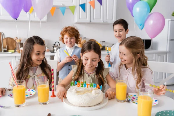 Chica feliz mirando su pastel de cumpleaños con velas cerca de amigos durante la celebración en casa - foto de stock