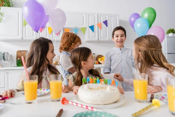 Niños alegres mirando feliz cumpleañero con frenos cerca de la torta con velas en la mesa - foto de stock
