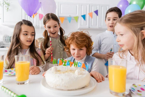 Pelirrojo chico soplando velas en el pastel de cumpleaños cerca de amigos durante la fiesta en casa - foto de stock
