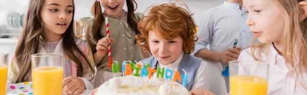 Ragazzo rosso che soffia candele sulla torta di compleanno vicino agli amici durante la festa a casa, banner — Foto stock