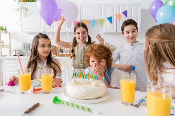 Рыжий мальчик прикрывает лицо, глядя на торт на день рождения рядом с друзьями во время вечеринки дома — стоковое фото