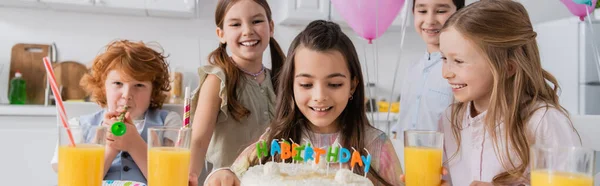 Menina alegre olhando para bolo de aniversário perto de amigos felizes durante a festa em casa, banner — Fotografia de Stock