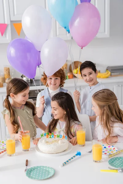 Niños alegres con globos mirando chica de cumpleaños cerca de la torta y cuernos de fiesta en la mesa - foto de stock