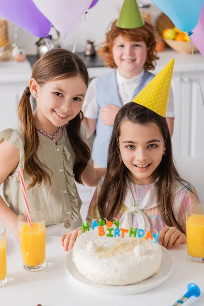 Happy girl in party cap looking at camera with friend near birthday cake — Stock Photo