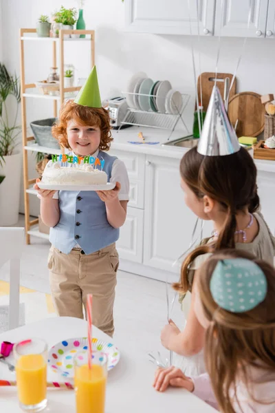 Menino ruiva feliz segurando bolo de aniversário com velas perto de amigos durante a festa em casa — Fotografia de Stock