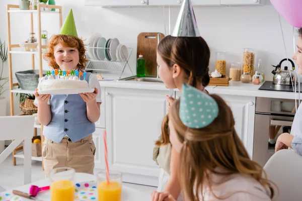Щасливий рудий хлопчик тримає торт на день народження зі свічками біля веселих друзів під час вечірки вдома — стокове фото