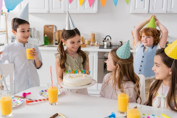 Glückliches Mädchen hält Geburtstagstorte mit Kerzen neben fröhlichen Freunden während der Party zu Hause — Stockfoto