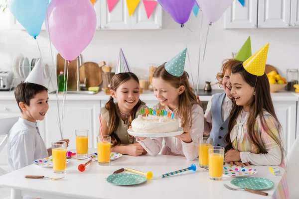 Happy girl holding birthday cake with candles near cheerful friends during celebration at home — Stock Photo