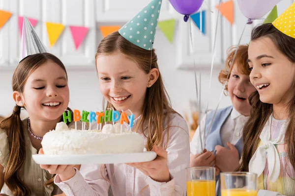 Chica feliz celebración de pastel de cumpleaños con velas cerca de niñas alegres durante la celebración en casa - foto de stock