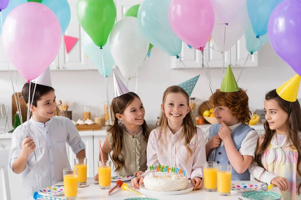 Grupo de niños felices en gorras de fiesta divirtiéndose durante la celebración de cumpleaños junto a globos en casa - foto de stock