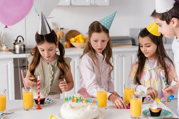 Preteen girls in party caps looking at birthday cake next to orange juice on table — Stock Photo