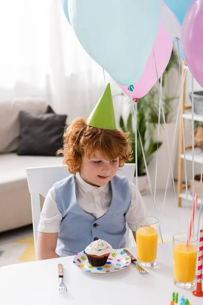 Curly redhead boy in party cap sitting next to glasses with orange juice and cupcake — Stock Photo