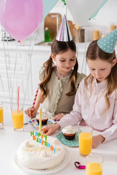 Preteen girls in party caps looking at birthday cake and cupcakes on table — Stock Photo