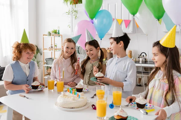 Grupo de niños alegres en gorras de fiesta divirtiéndose durante la fiesta de cumpleaños al lado de globos de colores - foto de stock