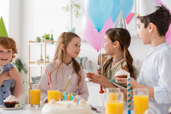 Happy girl looking at friend near boys in party caps during birthday party — Stock Photo