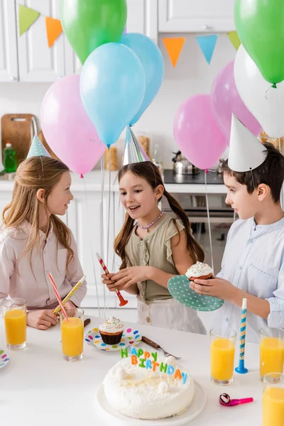 Preteen boy in party cap holding cupcake near cheerful girls next to balloons during birthday party — Stock Photo