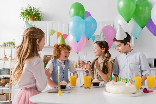 Счастливые девушки в шапочках для вечеринок хлопают в ладоши и поют счастливую песню дня рождения рядом с мальчиками и тортом — стоковое фото