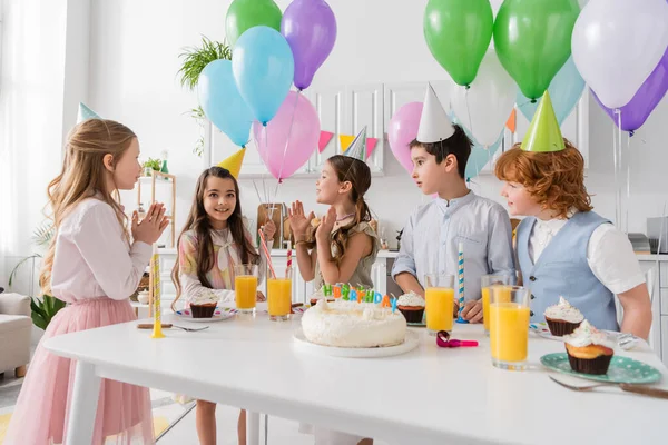 Niños felices aplaudiendo y cantando feliz cumpleaños canción al lado de la torta con velas y globos - foto de stock