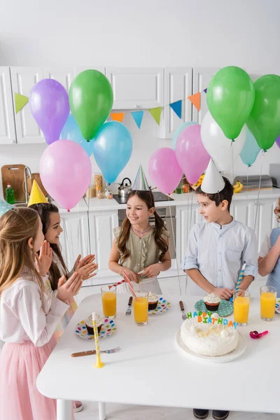 Niños felices aplaudiendo y cantando feliz cumpleaños al lado de la torta con velas y globos - foto de stock