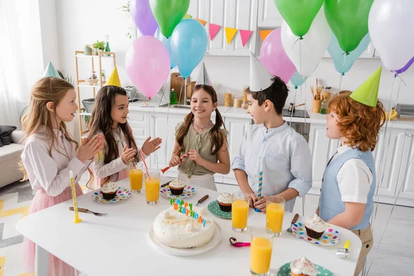 Положительные дети хлопают в ладоши и поют счастливую песню дня рождения рядом с тортом со свечами и воздушными шарами — стоковое фото