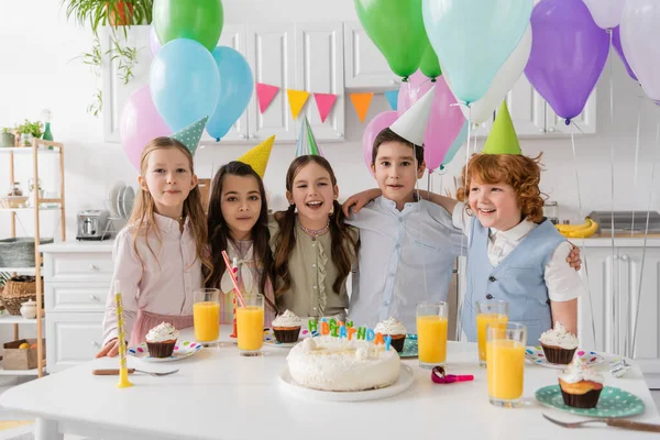 Grupo positivo de niños cantando feliz cumpleaños canción al lado de la torta con velas y globos - foto de stock