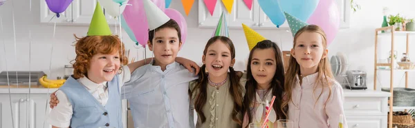 Grupo positivo de niños cantando feliz cumpleaños canción al lado de la torta con velas y globos, pancarta - foto de stock