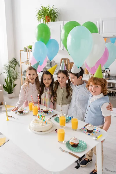 Grupo de niños felices cantando feliz canción de cumpleaños al lado de la torta con velas y globos - foto de stock