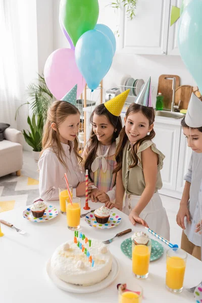 Happy preteen girls in party caps smiling next to cupcakes during birthday party — Stock Photo
