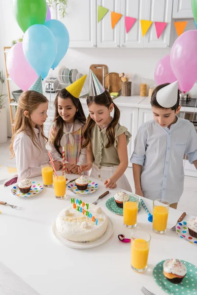 Happy preteen girls in party caps smiling next to boy and cupcakes during birthday party — Stock Photo