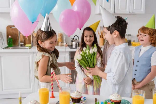 Preadolescente chico dando tulipanes a alegre cumpleaños chica cerca de amigos en fondo borroso - foto de stock