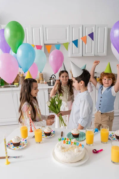Preteen boy giving flowers to cheerful birthday girl near friends on blurred background — Stock Photo