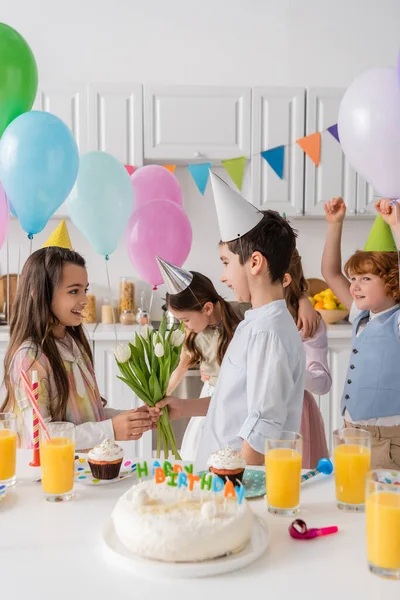 Preteen boy giving tulips to happy birthday girl near friends on blurred background — Stock Photo